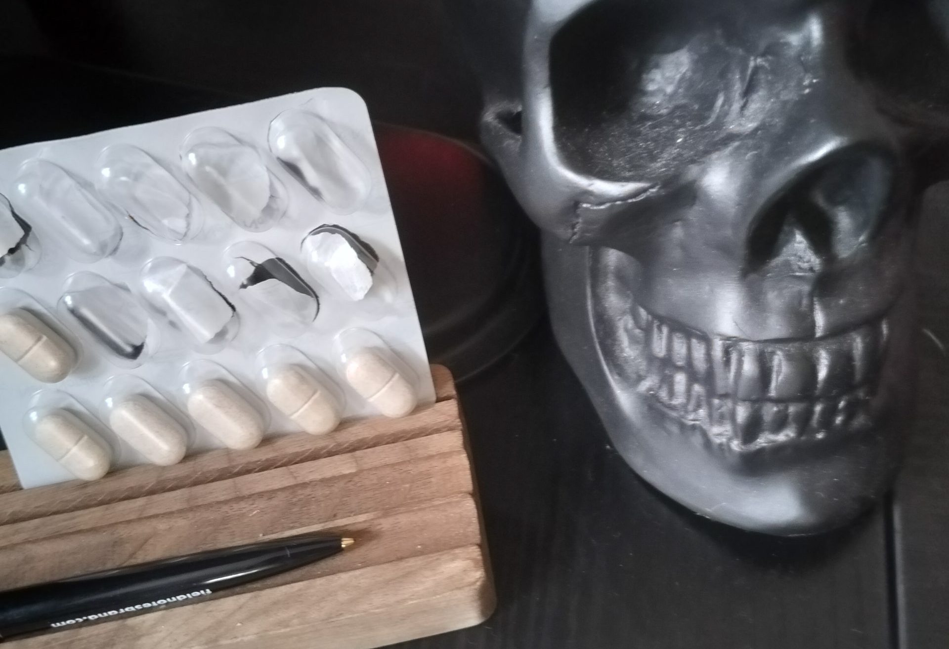 A blister pack of pills rests in a wooden pen stand, next to a black skull desk tidy on a black desk